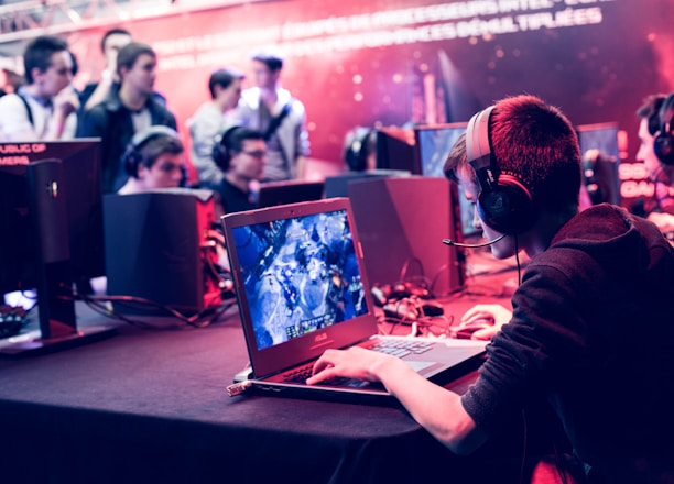 Several people are seated and focused on computer screens, wearing headsets, in an environment that appears to be a gaming tournament or esports event. The room is dimly lit with neon lighting, highlighting screens and creating an intense atmosphere.