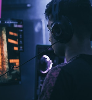 A person wearing headphones is engaged in gaming on a computer, with a large screen displaying a video game scene, showing a wooden building and a path.